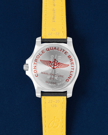Solid stainless steel screw-down caseback on a brand new Breitling Avenger GMT showing the yellow leather strap backing and red factory quality control sticker