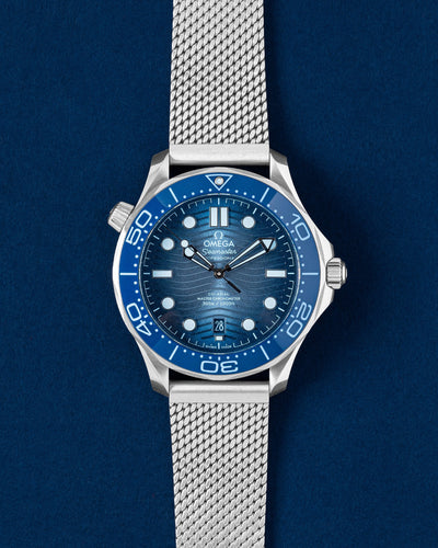A pre-owned 2024 75th Anniversary Omega Seamaster Diver 300M showcasing its gradient Summer Blue wave dial and matching blue ceramic bezel insert at Grand Caliber in Dallas