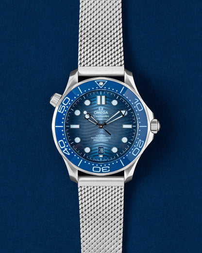 A pre-owned 2024 75th Anniversary Omega Seamaster Diver 300M showcasing its gradient Summer Blue wave dial and matching blue ceramic bezel insert at Grand Caliber in Dallas