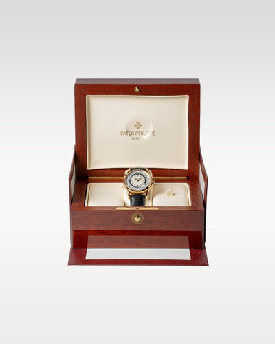 18k rose gold Patek Philippe World Time watch resting upright on its cushion inside its open high-gloss lacquered luxury wooden presentation box
