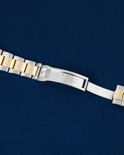 flat top-down view of the internal folding clasp blades and folding mechanism of a yellow gold and steel Rolex Oyster bracelet open over a navy blue background