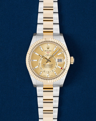 2021 Rolex Sky-Dweller 326933 watch featuring a champagne sunray dial, 18k yellow gold fluted Ring Command bezel and two-tone Oyster bracelet on a navy blue studio background