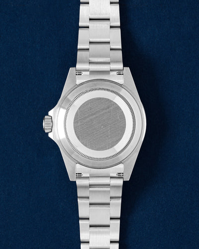 Rear studio portrait view showing the brushed stainless steel screw-down caseback, Oyster bracelet links, and case flanks flat-laid against a blue surface