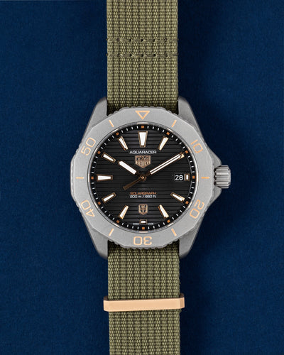 40mm titanium TAG Heuer Aquaracer Professional 200 Solargraph Sundowner watch featuring a black dial, rose gold bezel accents, and a khaki green NATO strap resting on a navy blue background