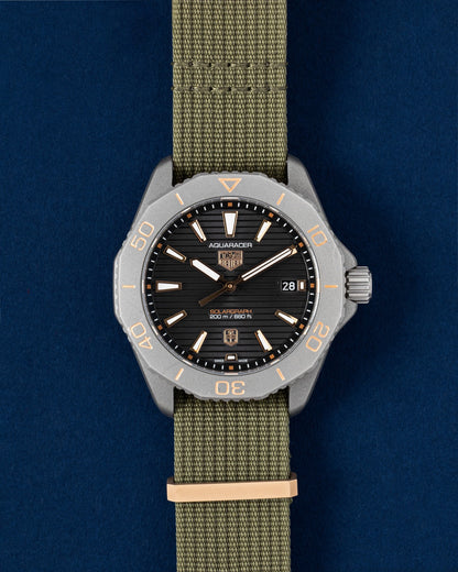 40mm titanium TAG Heuer Aquaracer Professional 200 Solargraph Sundowner watch featuring a black dial, rose gold bezel accents, and a khaki green NATO strap resting on a navy blue background
