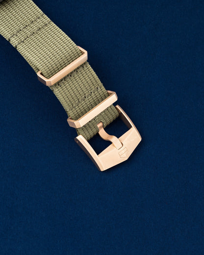 angled studio close-up of the rose gold-plated titanium tang buckle and fabric loop on the khaki green TAG Heuer NATO strap over a navy blue backdrop