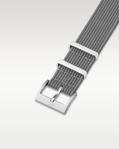 Angled detail shot of a charcoal grey nylon watch band showing its pre-installed brushed silver pin buckle