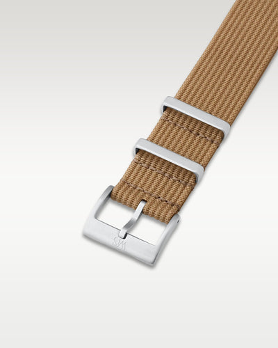 Angled detail shot of a sandy desert tan nylon watch band showing its pre-installed brushed silver pin buckle