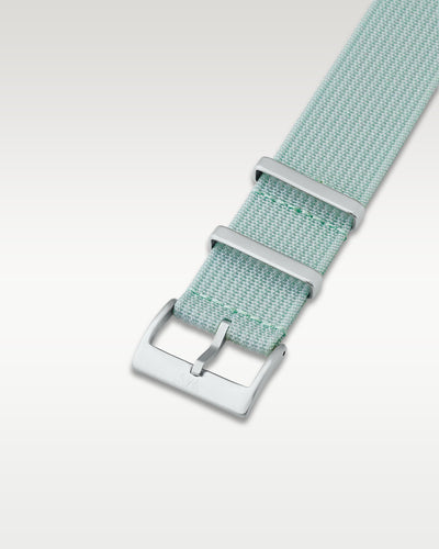 Angled detail shot of a subtle sage green nylon watch band showing its pre-installed brushed silver pin buckle